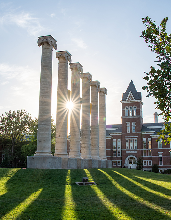 Sunlight shining through six stone columns on a summer evening with a brick building in the background.